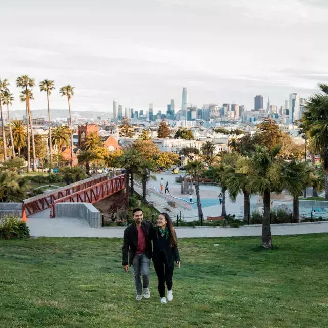 Una pareja camina hacia la cámara con Dolores Park y el Skyline 贝博体彩app detrás de ellos.